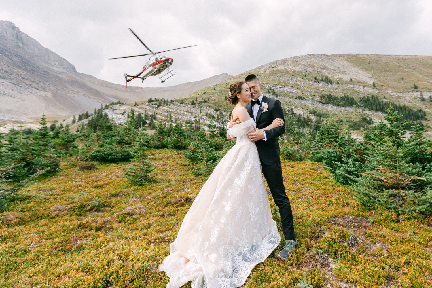 A couple embraces in a stunning mountain setting while a helicopter flies overhead, capturing the essence of their unique outdoor wedding.