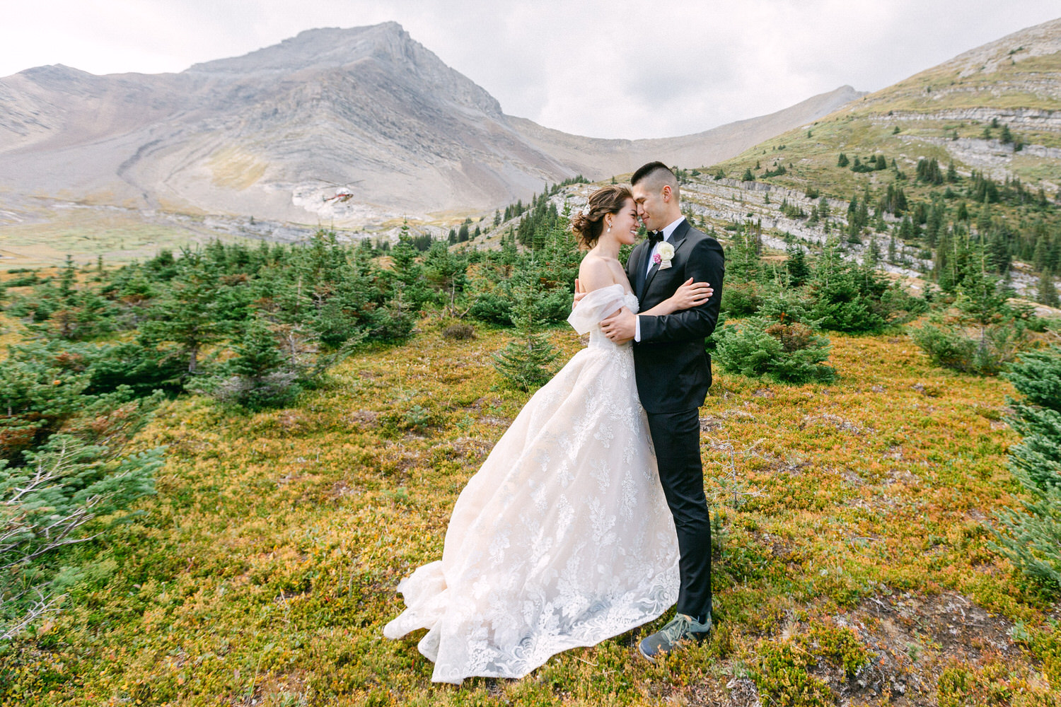 A couple in formal attire embracing in a scenic outdoor setting surrounded by lush greenery and mountains.