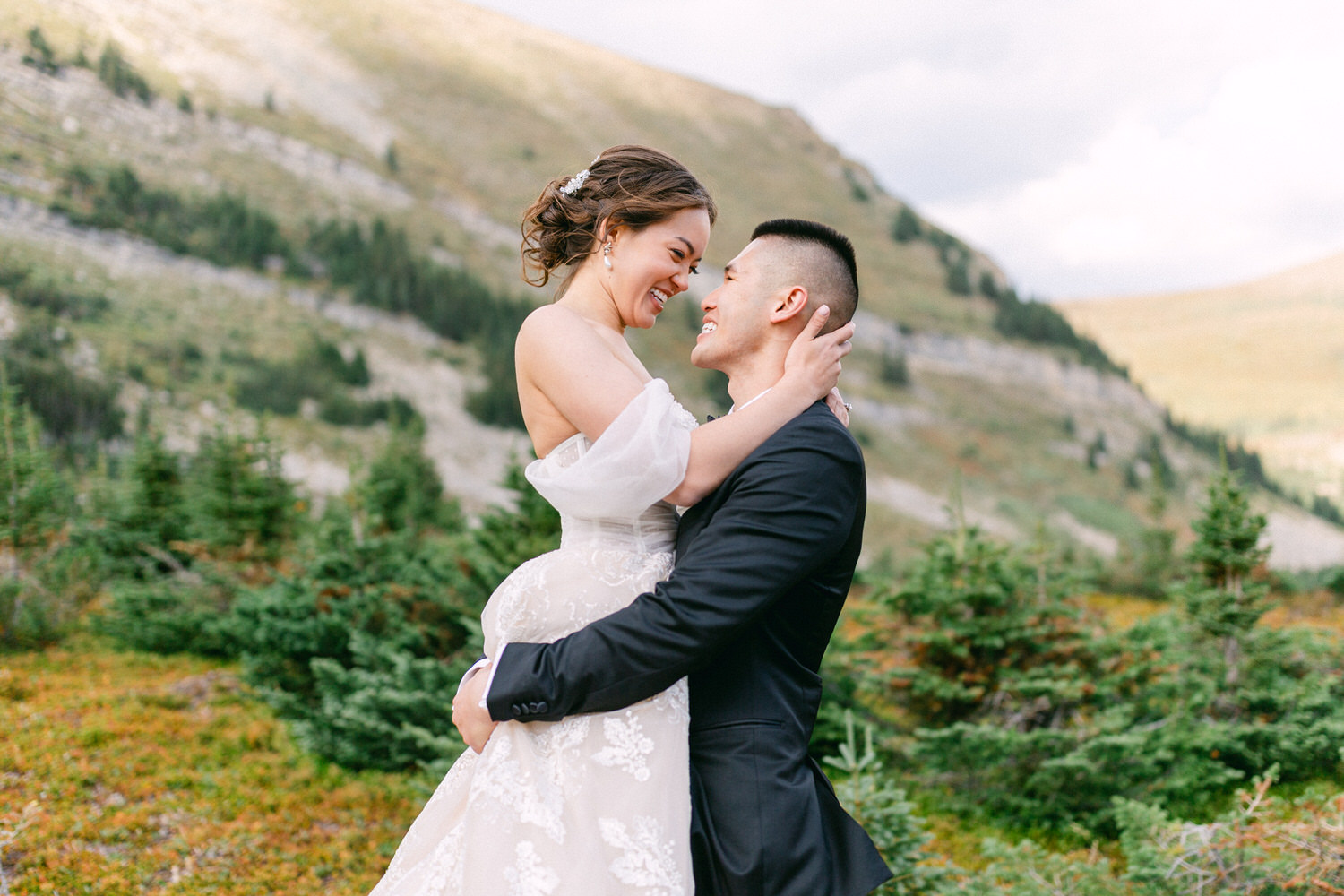 A bride and groom share a joyful moment in a picturesque mountain landscape, with the bride elegantly dressed in a white gown and the groom in a black suit.