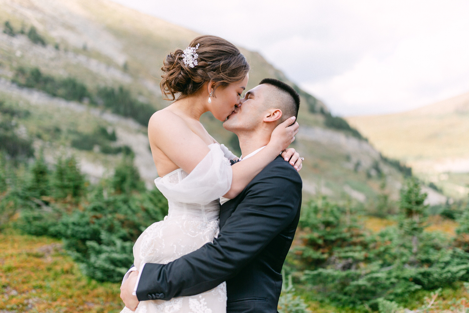 A couple shares a passionate kiss in a stunning mountain landscape, surrounded by lush greenery and rocky hills.