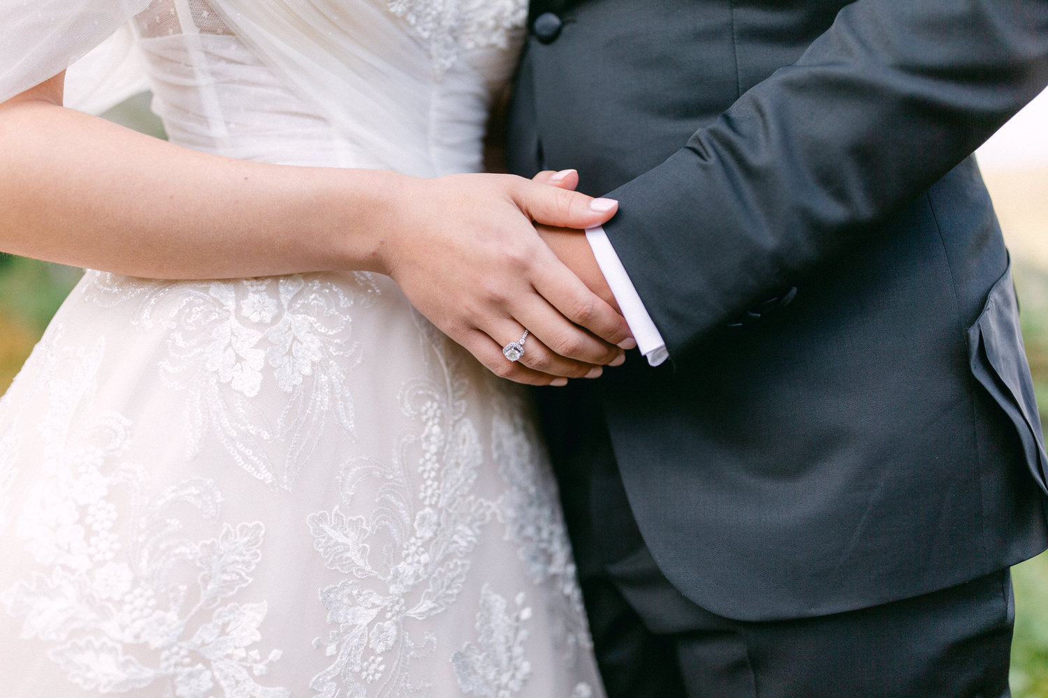 Close-up of a bride's and groom's hands clasped together, showcasing a diamond engagement ring and elegant wedding attire.