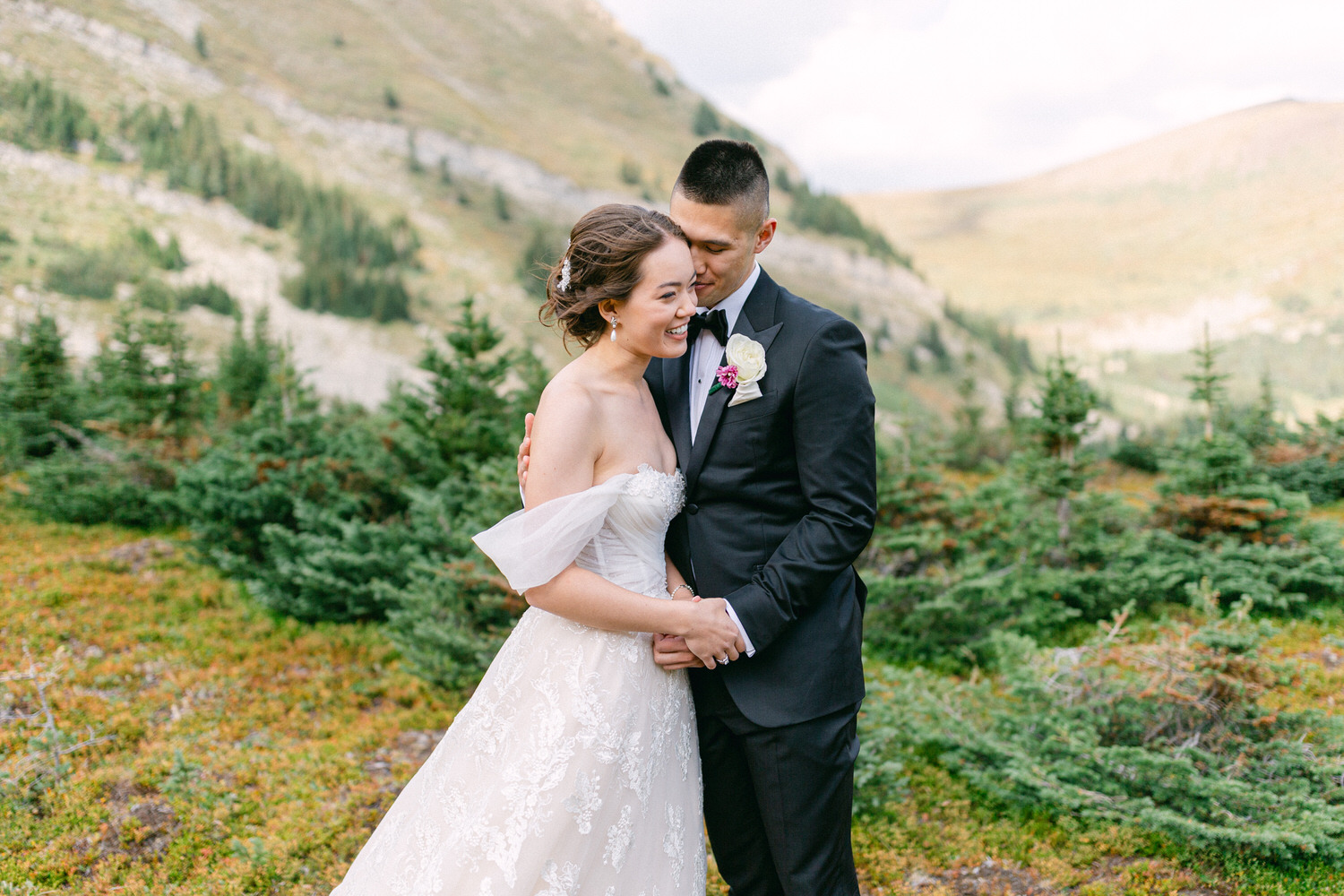 A happy couple embraces in a scenic outdoor setting, surrounded by lush greenery and mountains, showcasing their love in a beautiful wedding attire.