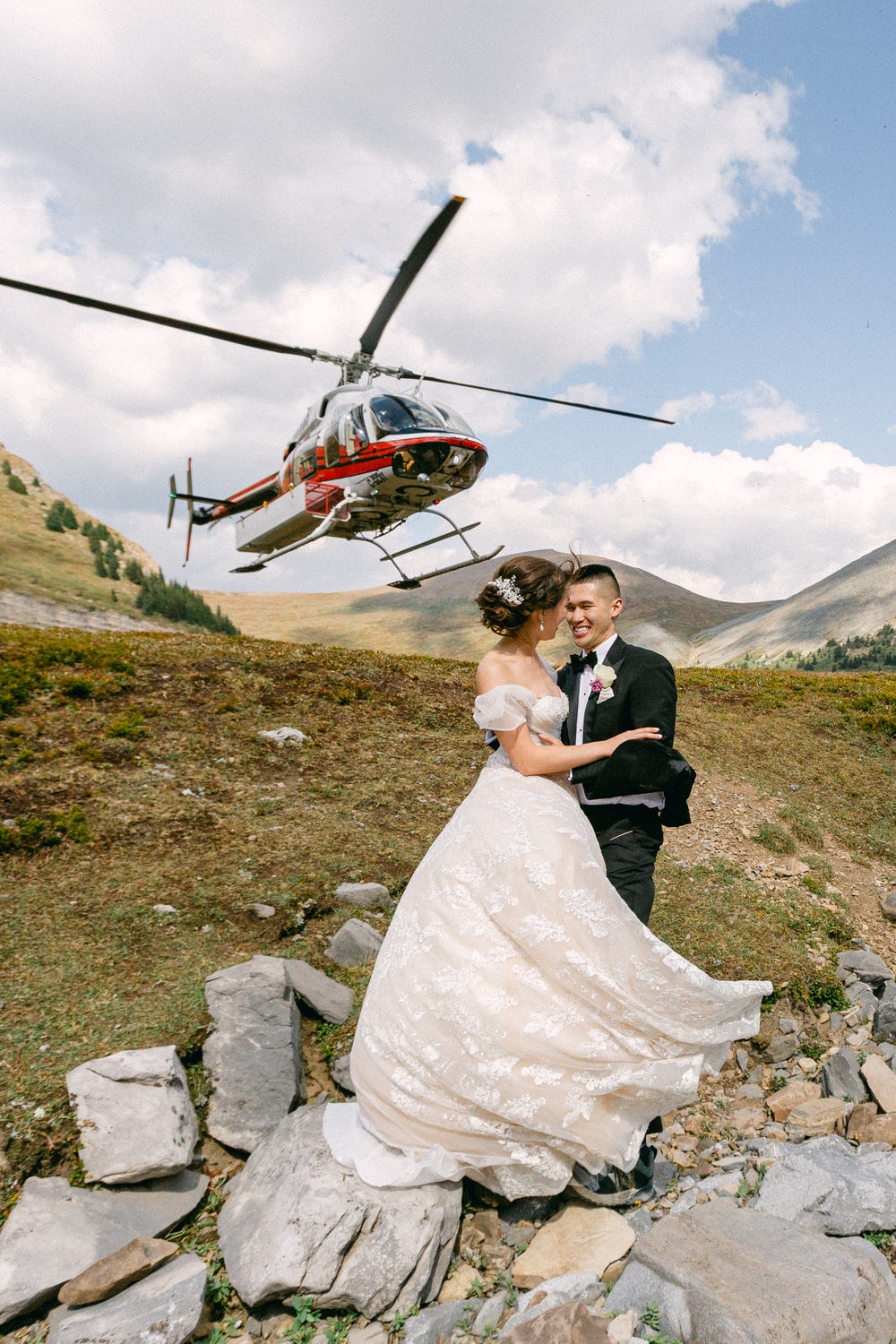 A joyful couple in wedding attire embraces outdoors, with a helicopter flying overhead amidst a picturesque mountain backdrop.