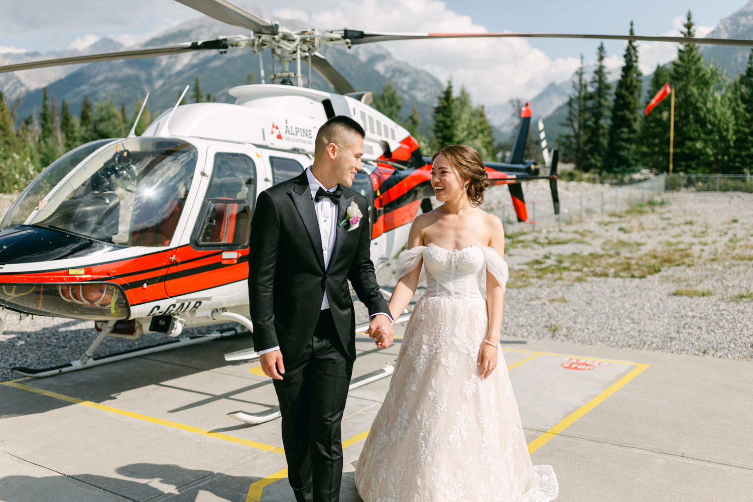 A happy bride and groom hold hands and smile at each other near a helicopter in a picturesque mountain setting.