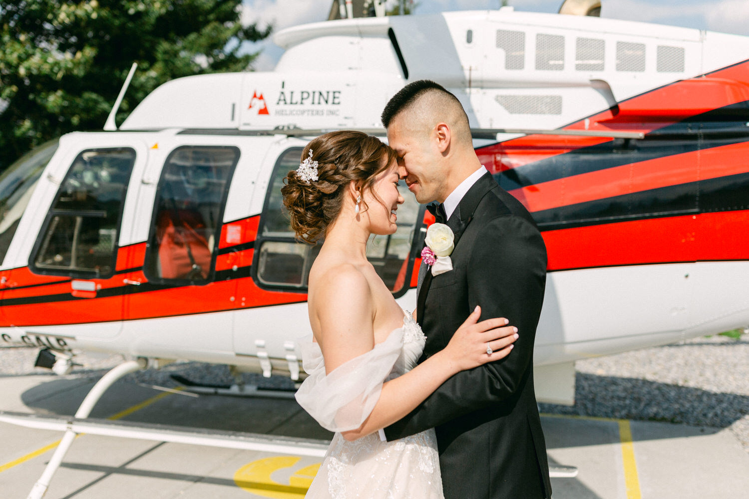 A bride and groom embrace tenderly in front of a red and white helicopter, celebrating their love on a sunny day.