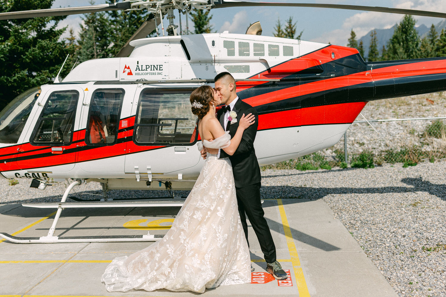 A couple shares a romantic kiss in front of a red and white helicopter, capturing a moment of love and adventure on their special day.