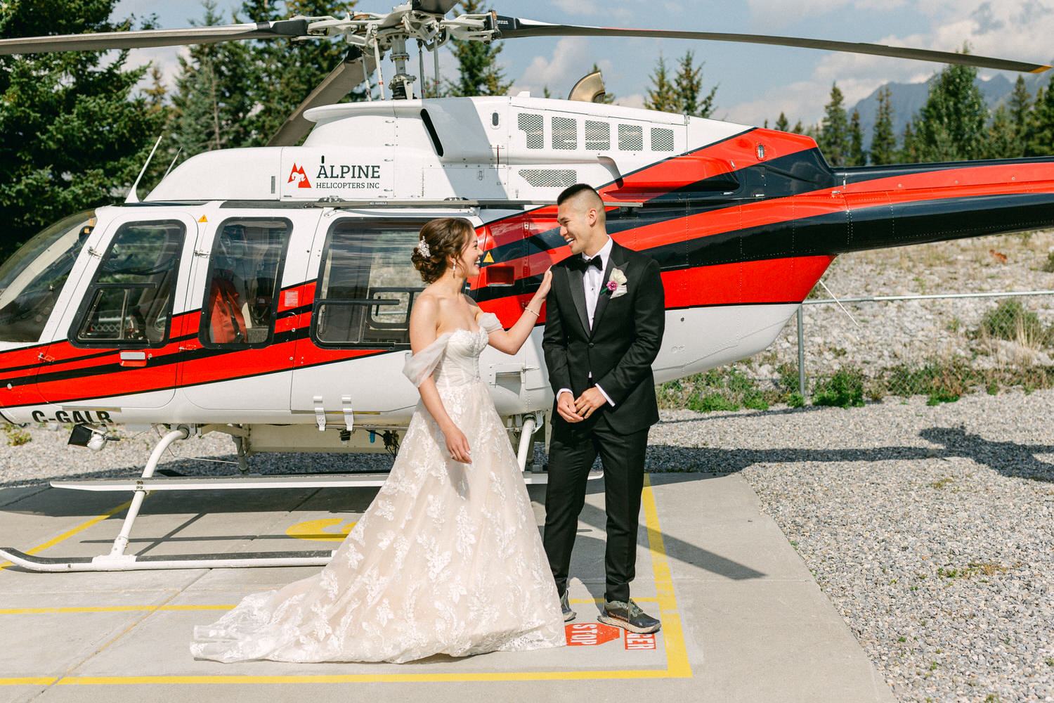 A bride and groom share a joyful moment in front of a striking red and white helicopter, capturing the excitement of their special day.