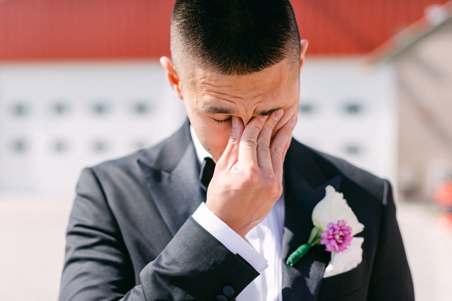 A man in a tuxedo wipes his brow, visibly emotional, with a flower boutonnière on his lapel.