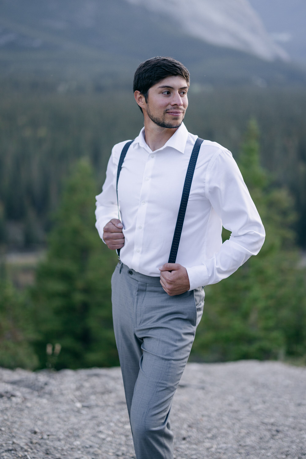 A well-dressed man in a white shirt and gray pants with suspenders stands confidently against a scenic mountainous backdrop.