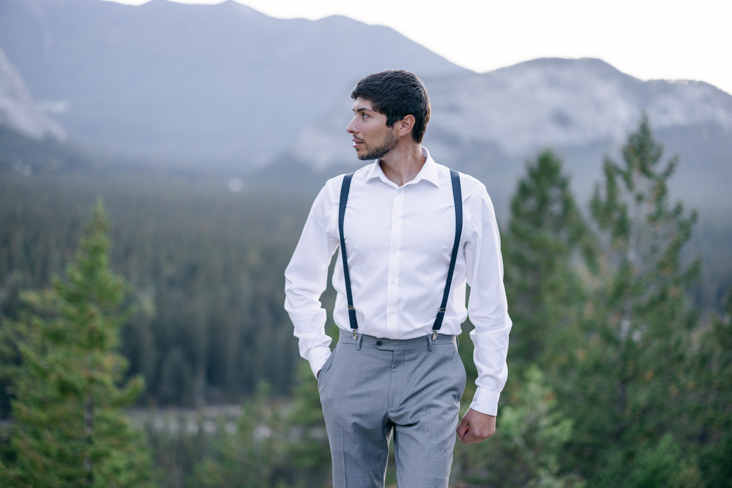 A well-dressed man in a white shirt and gray trousers with suspenders stands against a backdrop of evergreen trees and mountains.