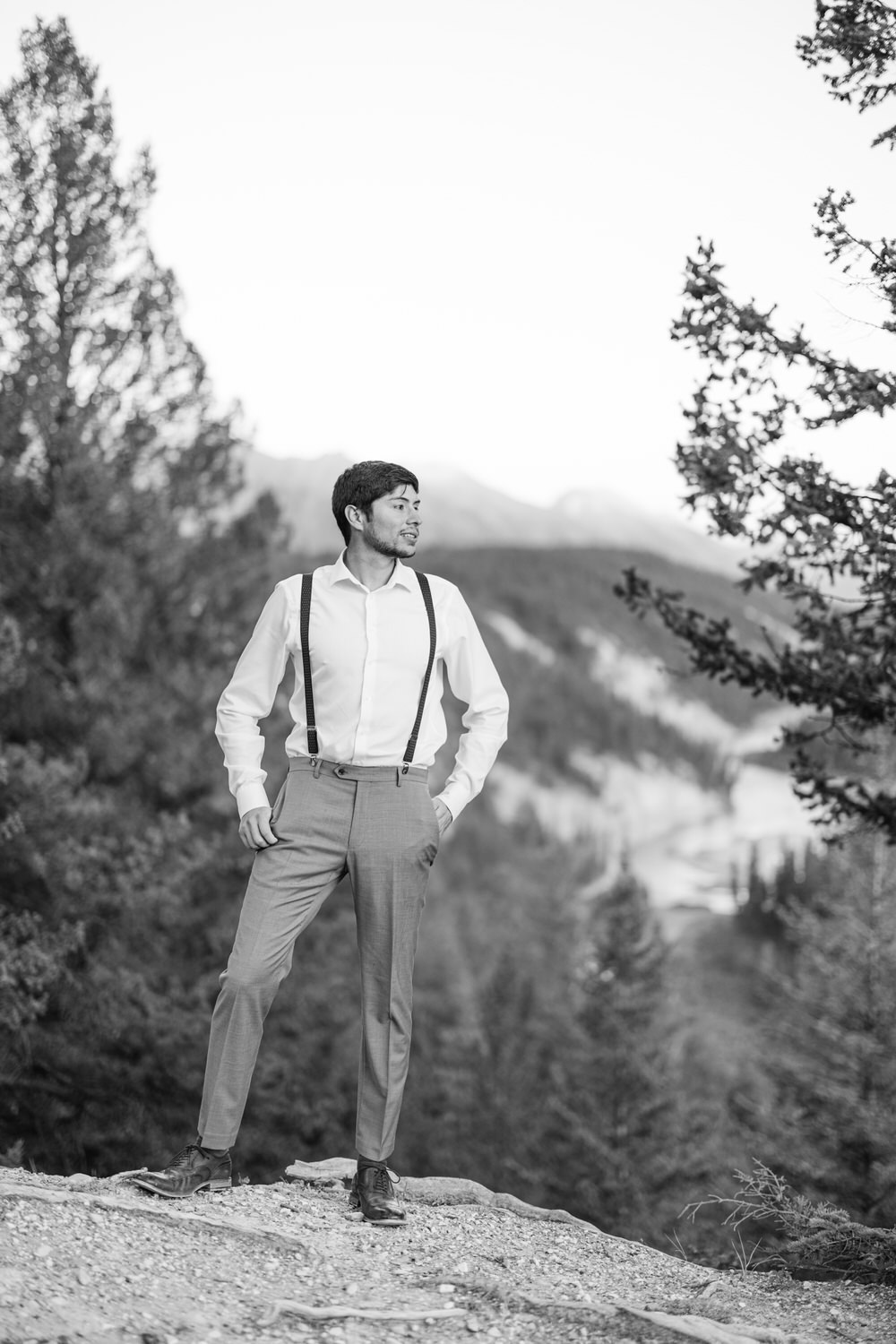 Elegant Man in Nature::A well-dressed man in suspenders poses confidently amidst a scenic outdoor landscape, featuring trees and mountains in the background.