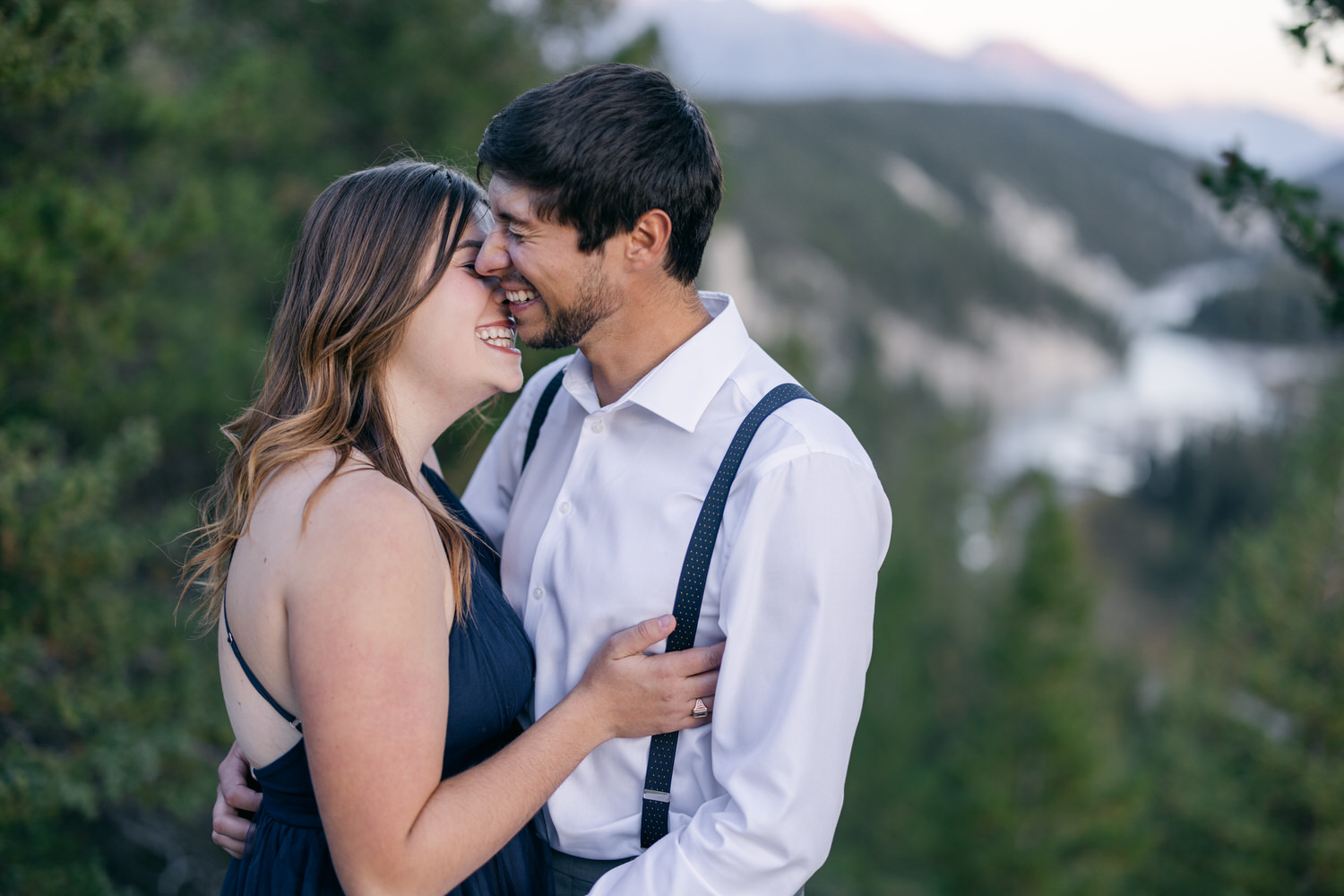 A couple shares a tender moment, smiling and leaning closer together against a beautiful forest backdrop.