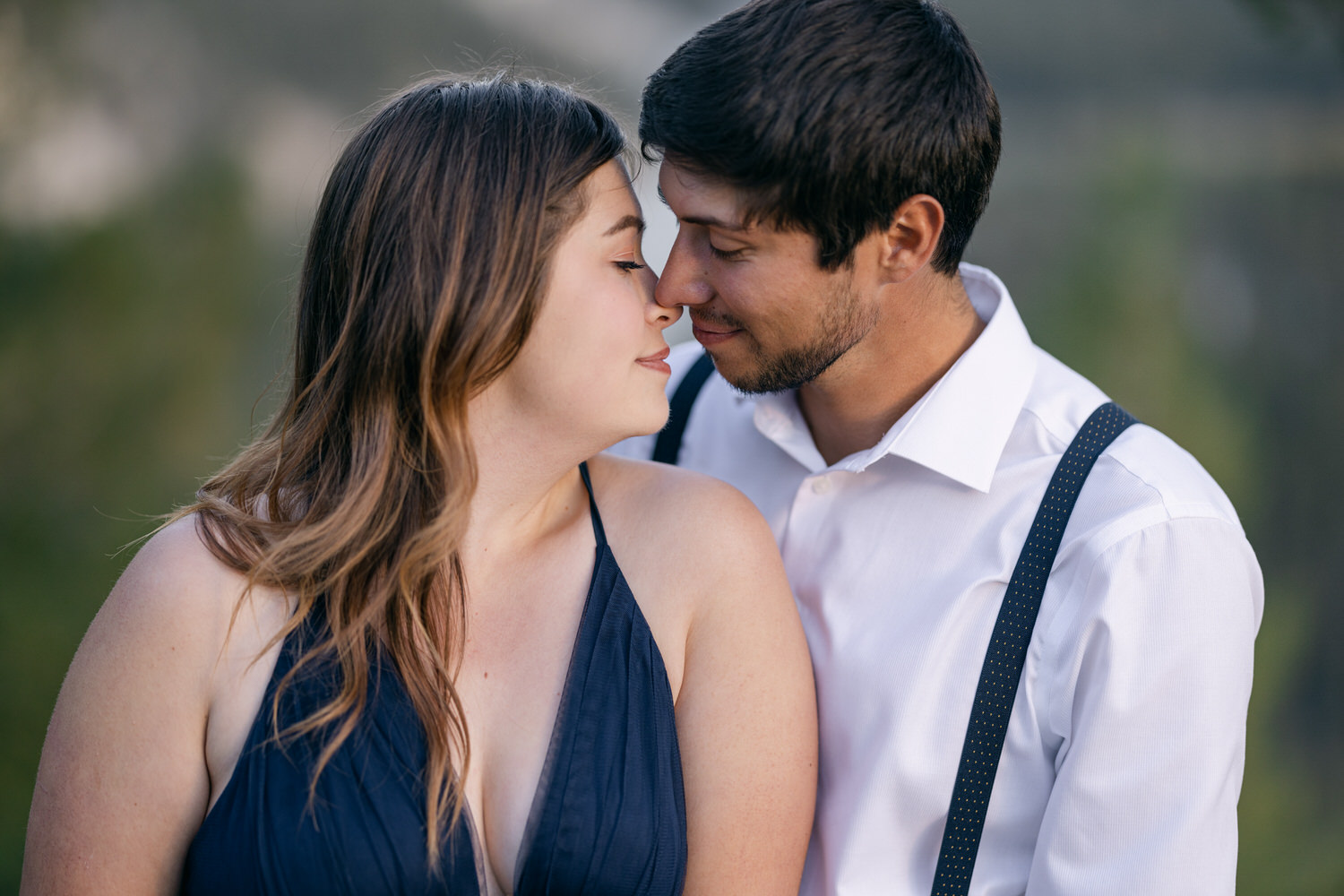 A close-up of a couple sharing a tender moment, leaning in for a nose-to-nose connection, surrounded by a blurred natural background.