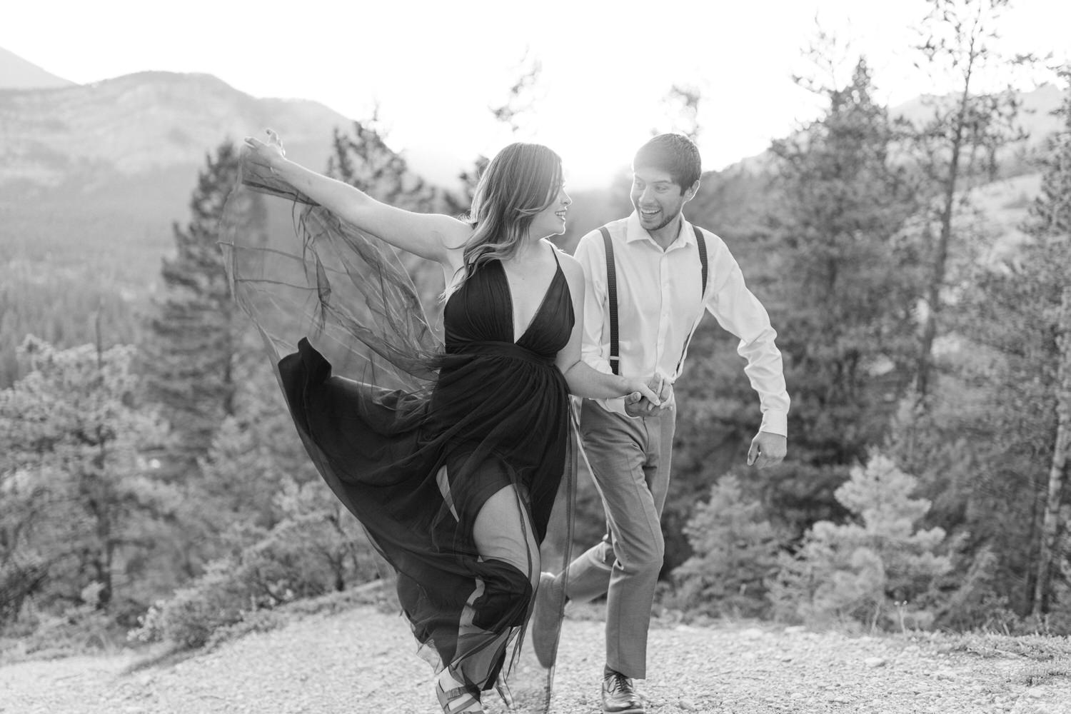 A couple joyfully running hand in hand through a scenic landscape, with mountains and trees in the background, captured in black and white.