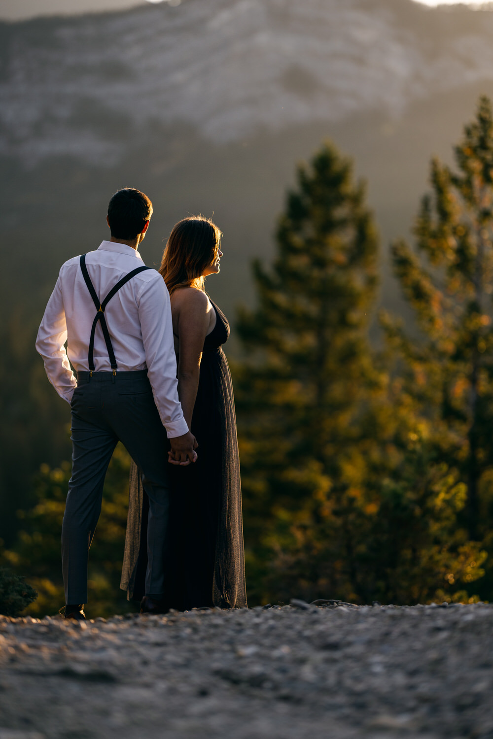 A couple stands hand in hand, silhouetted against a sunset backdrop, surrounded by nature.