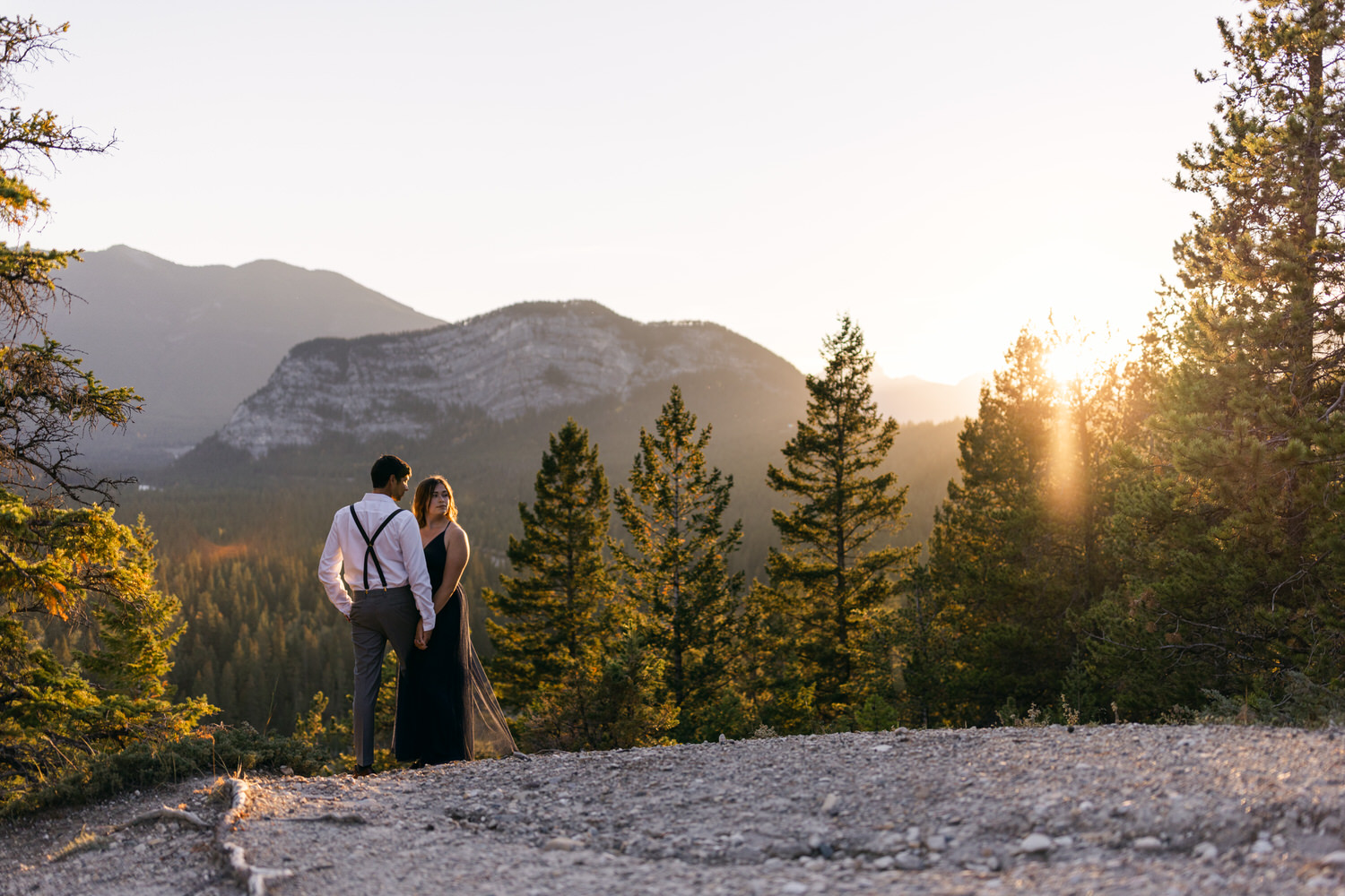 A couple holding hands in a scenic mountain landscape during sunset, surrounded by tall trees.