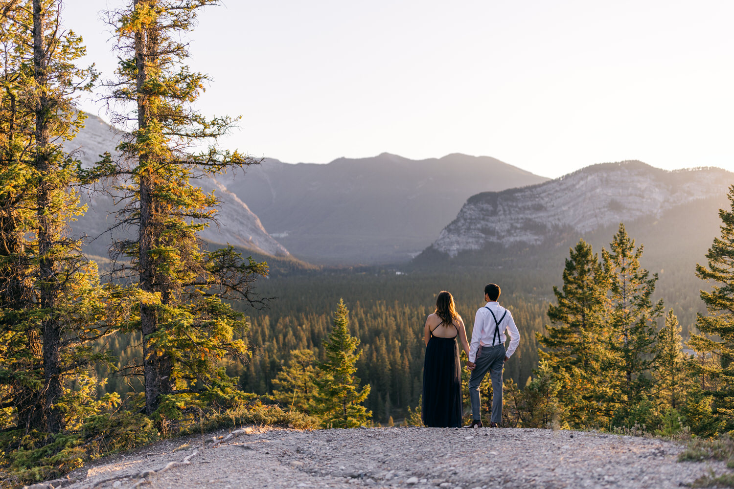 A couple stands hand in hand, gazing at a breathtaking mountainous landscape illuminated by the warm glow of sunset, surrounded by lush trees.