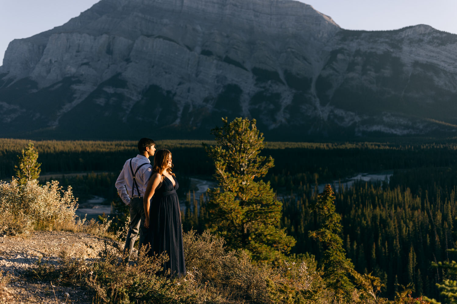 A couple stands on a rocky ledge overlooking a vast forest and mountains, bathed in golden sunlight during the evening.