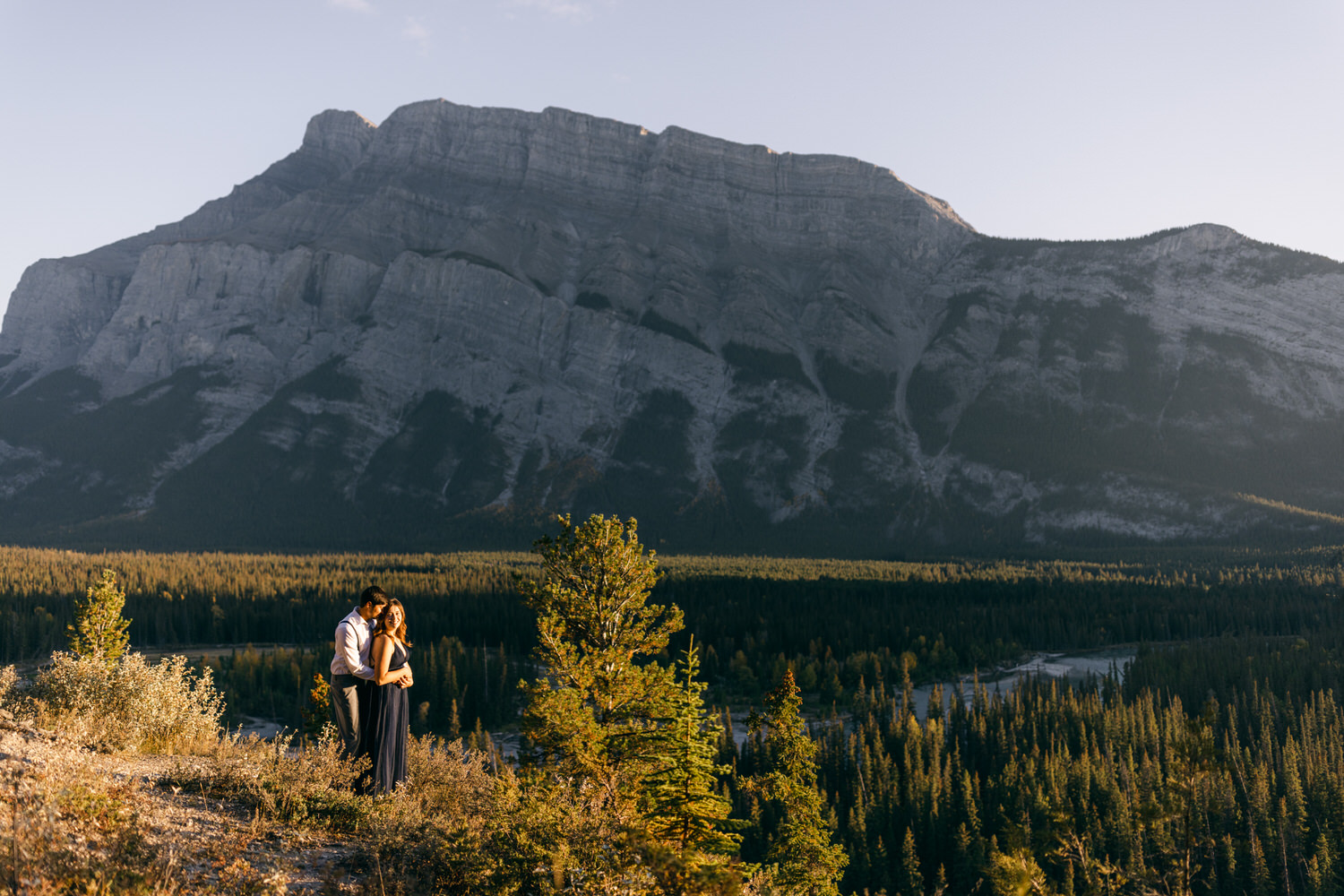 A couple embraces on a rocky outcrop with a majestic mountain backdrop during sunset.