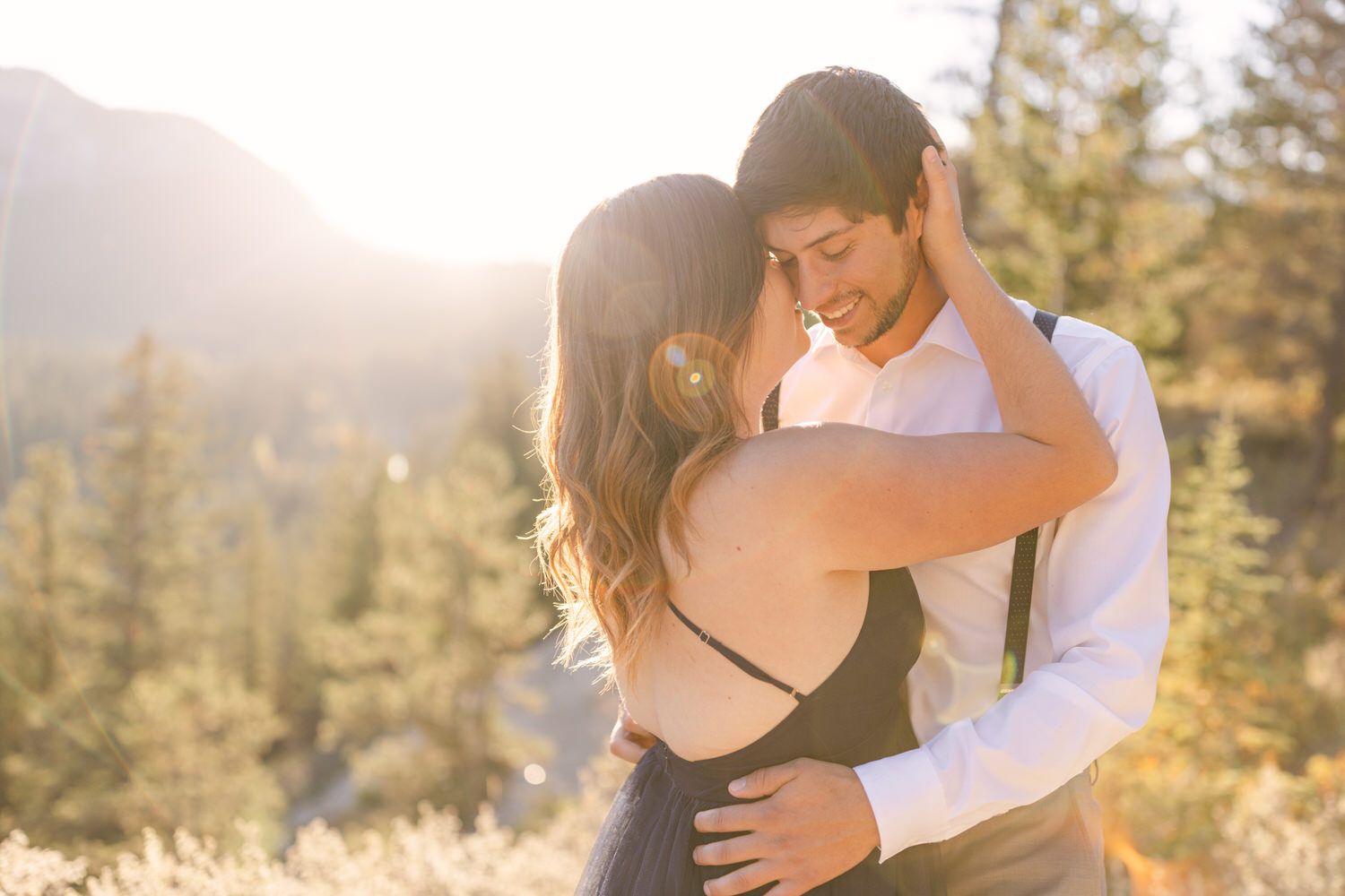 A couple share a tender moment in a sunlit outdoor setting, surrounded by trees and mountains, highlighting their connection and affection.