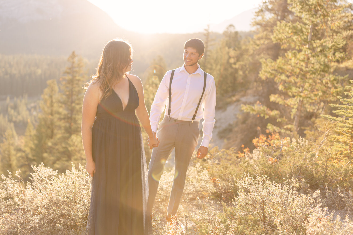 A couple holds hands in a sunlit outdoor setting, surrounded by trees and mountains, radiating warmth and romance.