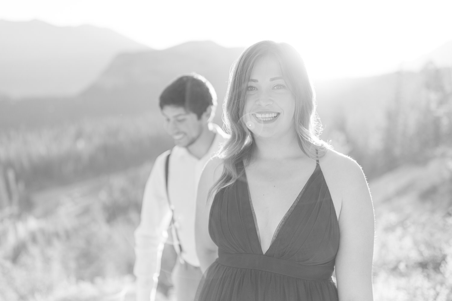 A smiling woman in a dark dress stands in the foreground, while a man in suspenders walks behind her, set against a scenic backdrop of mountains and soft sunlight.