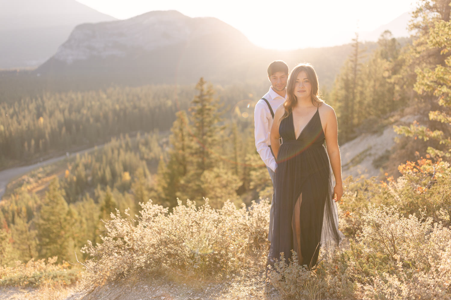 A couple stands together on a hillside during sunset, with a backdrop of mountains and trees. The woman, in a flowing dark dress, walks forward while the man stands behind her, both bathed in warm golden light.