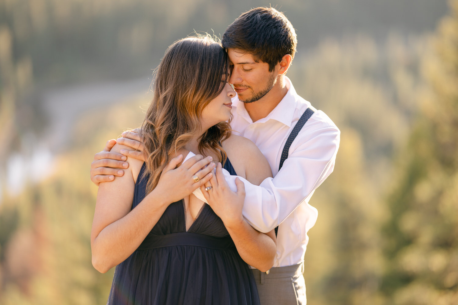 A man and woman share a tender moment outdoors, with the woman in a dark dress and the man in a white shirt and suspenders, surrounded by a blurred natural background.