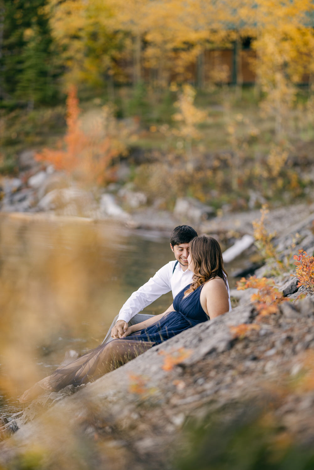 A couple sitting on rocky shore, gazing at each other amidst vibrant autumn foliage by the water.