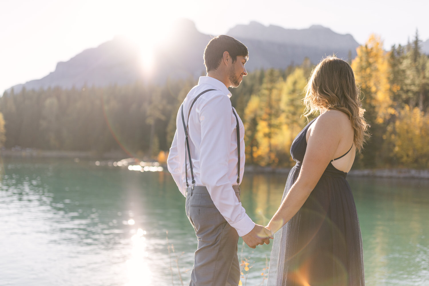 A couple stands hand in hand by a tranquil lake, surrounded by autumn foliage and mountains, capturing a moment of connection in golden sunlight.