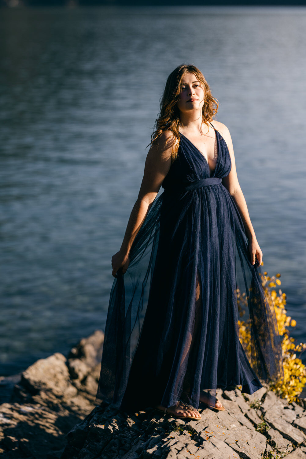 A woman in a flowing navy blue dress stands on rocky terrain beside a serene body of water, exuding confidence and grace.