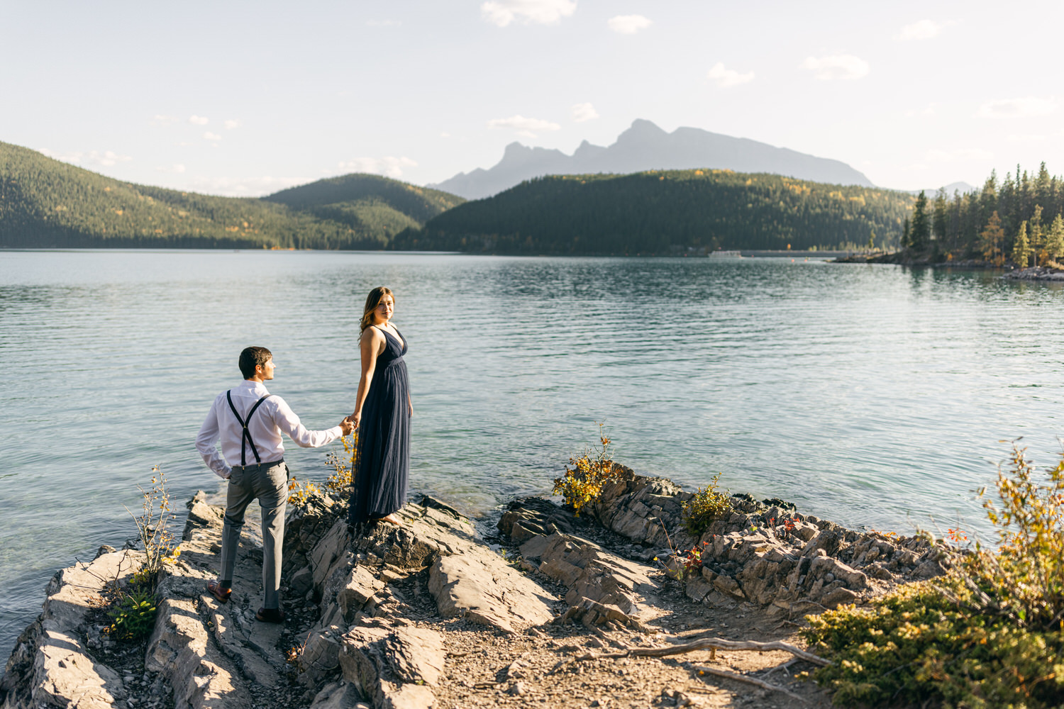 Serene Lake Engagement::A couple stands on rocky shore, holding hands with mountains and reflection in the background.