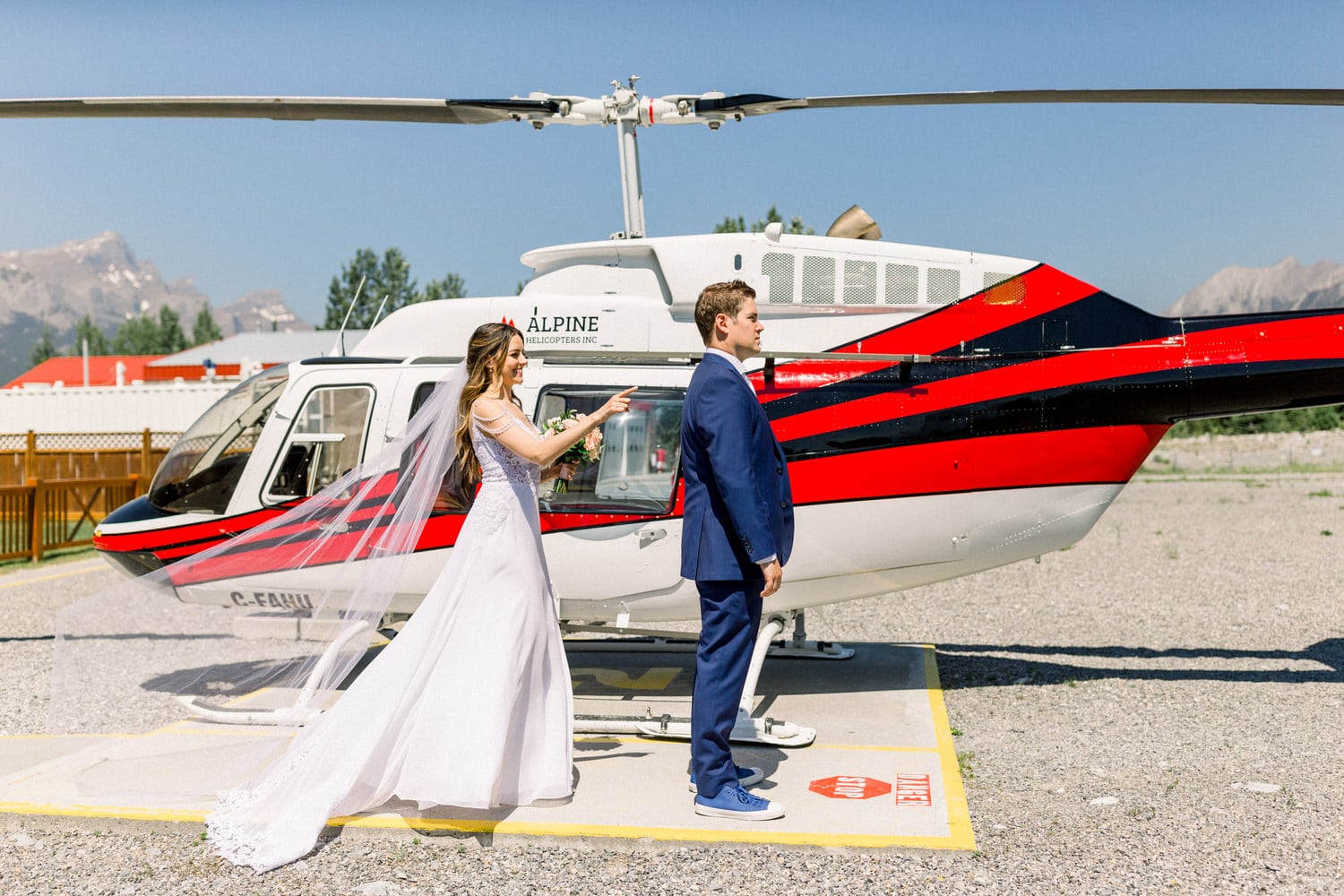 A bride in a flowing white gown surprises her groom next to a red and black helicopter on a sunny day.