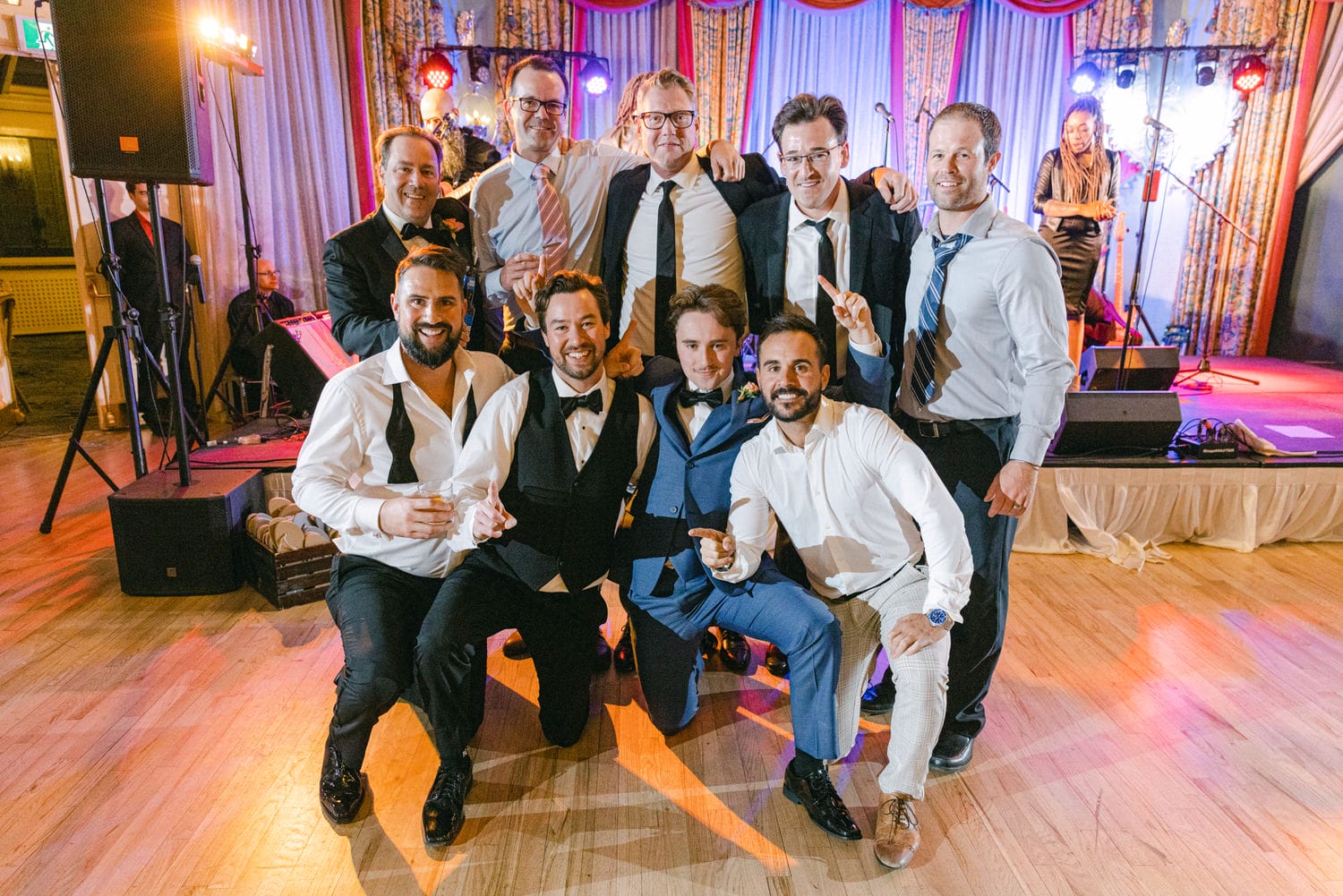 A group of well-dressed men joyfully posing together at a lively event, with a stage and musical equipment in the background.