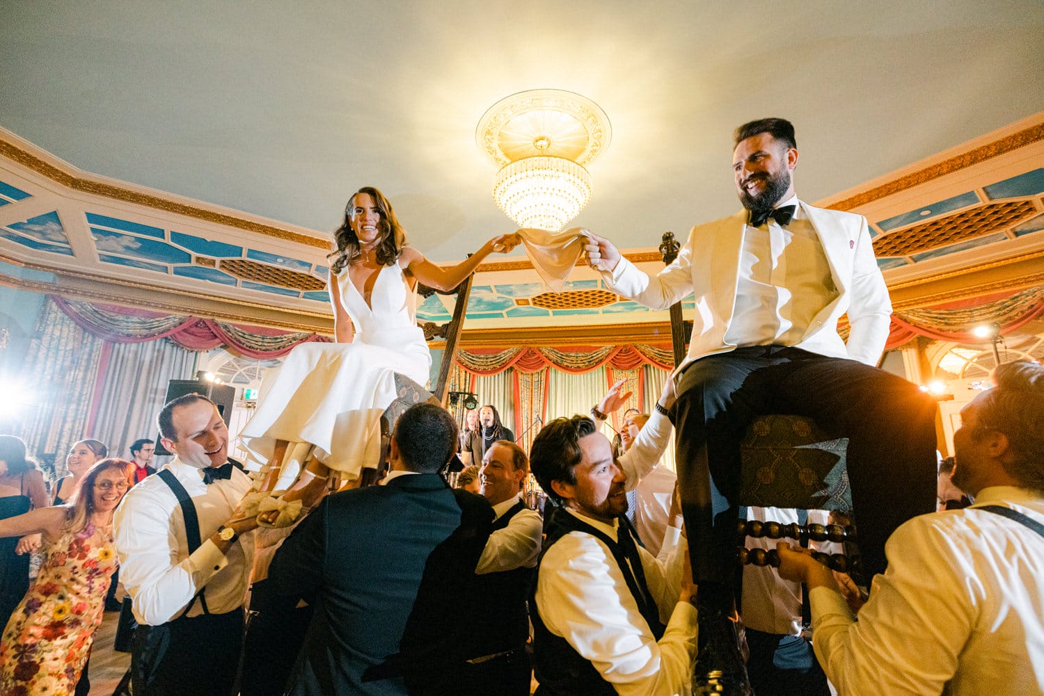 A joyful moment capturing the bride and groom being lifted in chairs by their friends during a celebratory wedding dance, surrounded by guests in a decorated ballroom.