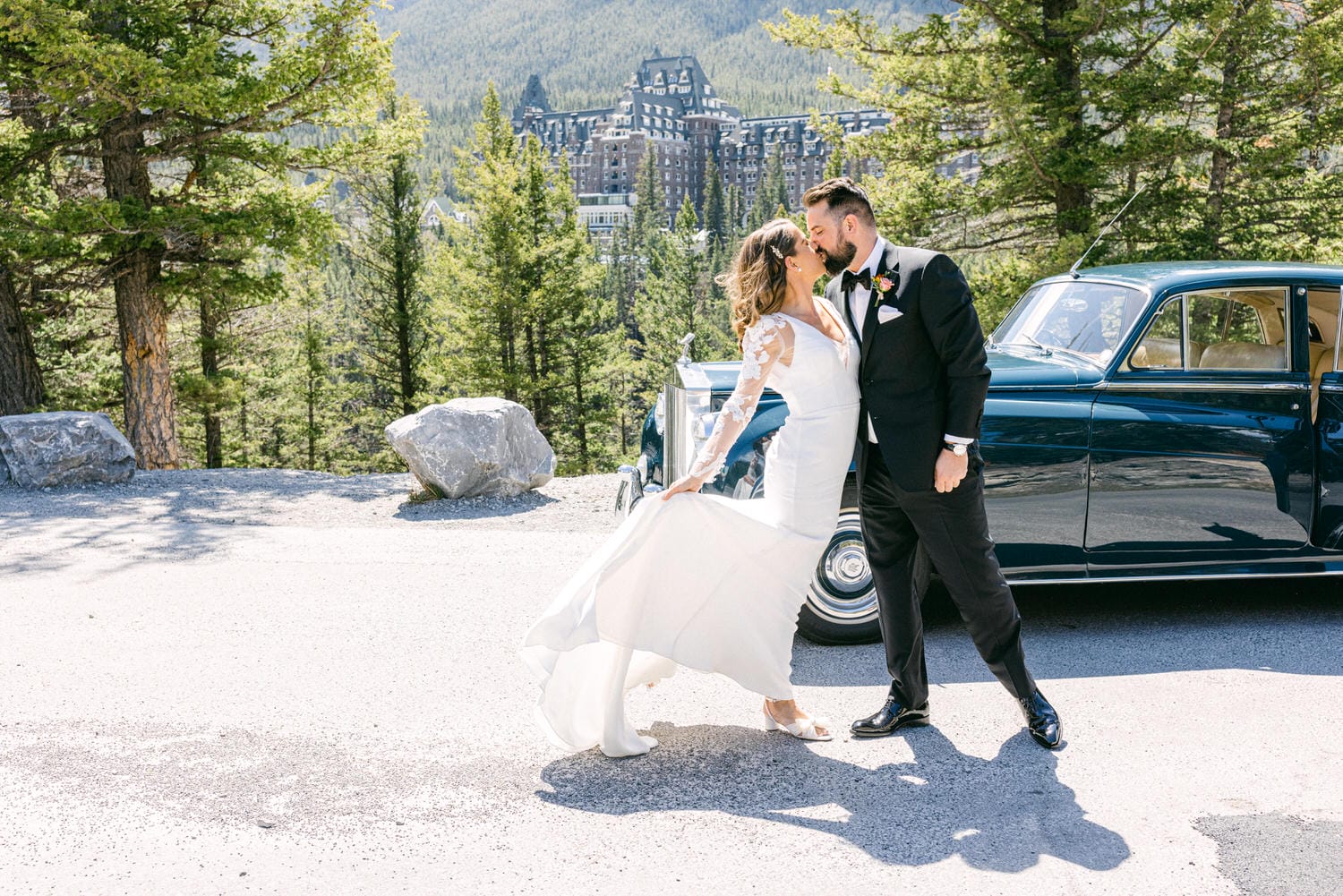 Wedding Bliss in Nature::A couple sharing a romantic kiss surrounded by trees and a classic vintage car, with a majestic building in the background.