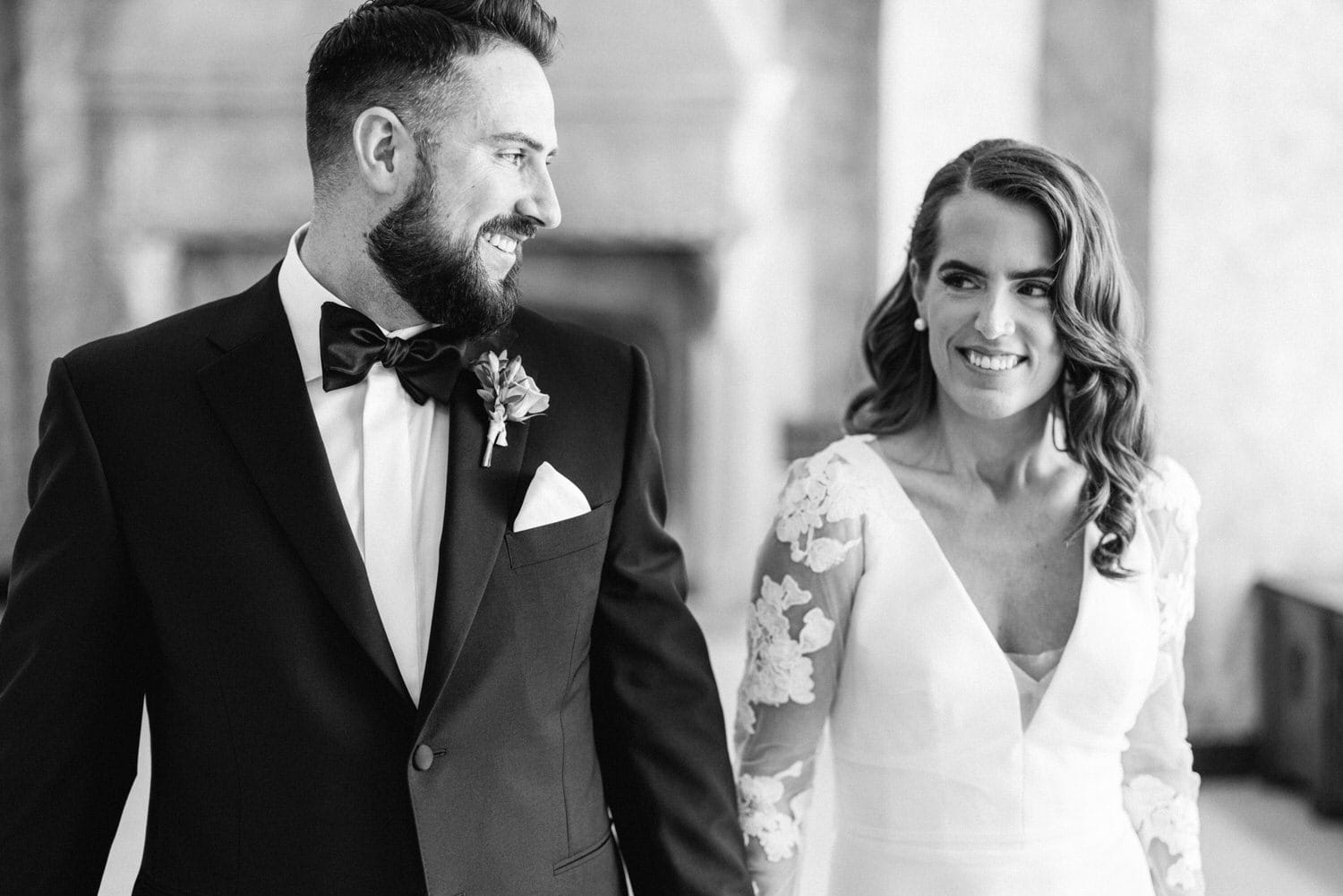 A groom in a tuxedo smiles at his bride, who wears a long-sleeved wedding dress with lace details, as they walk hand in hand in a beautiful venue.