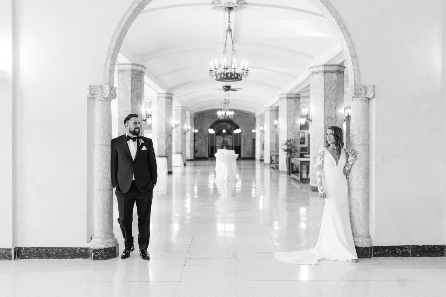 A bride and groom are poised in a grand hall, with a stunning architectural backdrop, showcasing their classic wedding attire in a romantic setting.