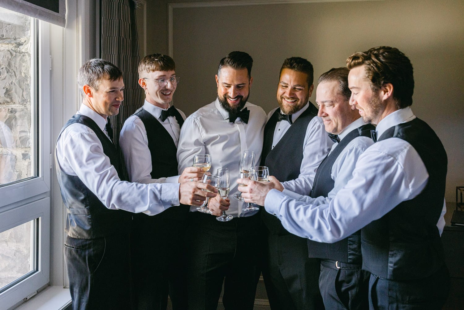 A group of well-dressed groomsmen toasting with glasses in a bright room, exuding joy and camaraderie.