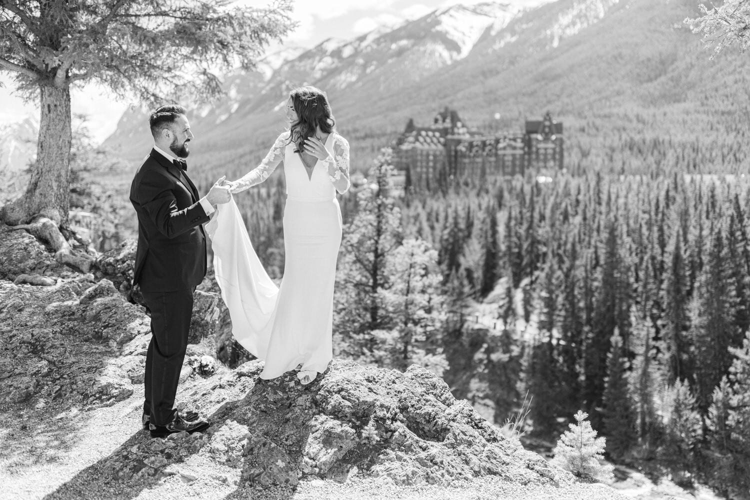 A couple shares a romantic moment on a rocky outcrop, with a breathtaking mountain backdrop and a historic hotel in the distance.