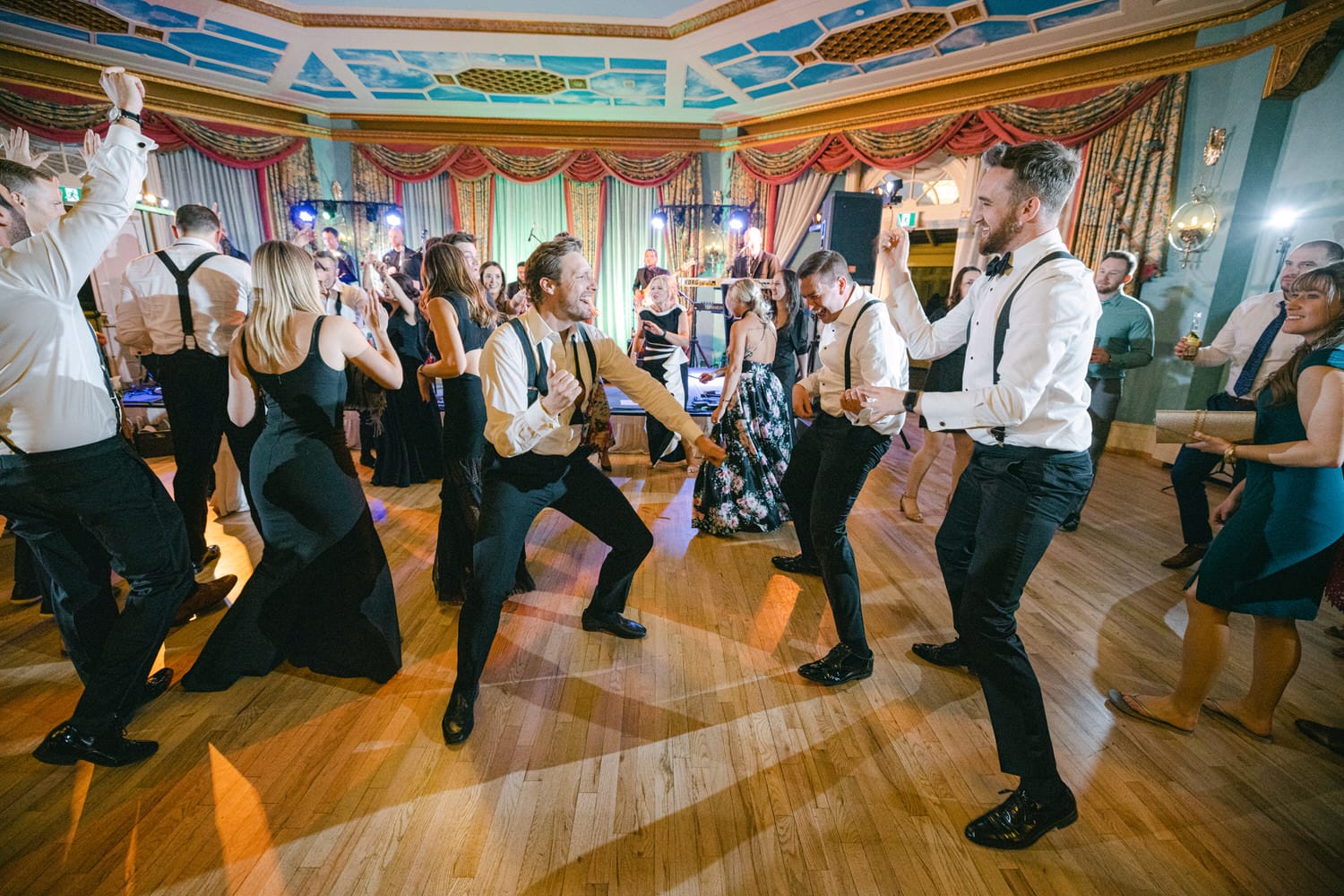 Guests in formal attire joyfully dancing at an elegant event, with a lively atmosphere and colorful lights in the background.