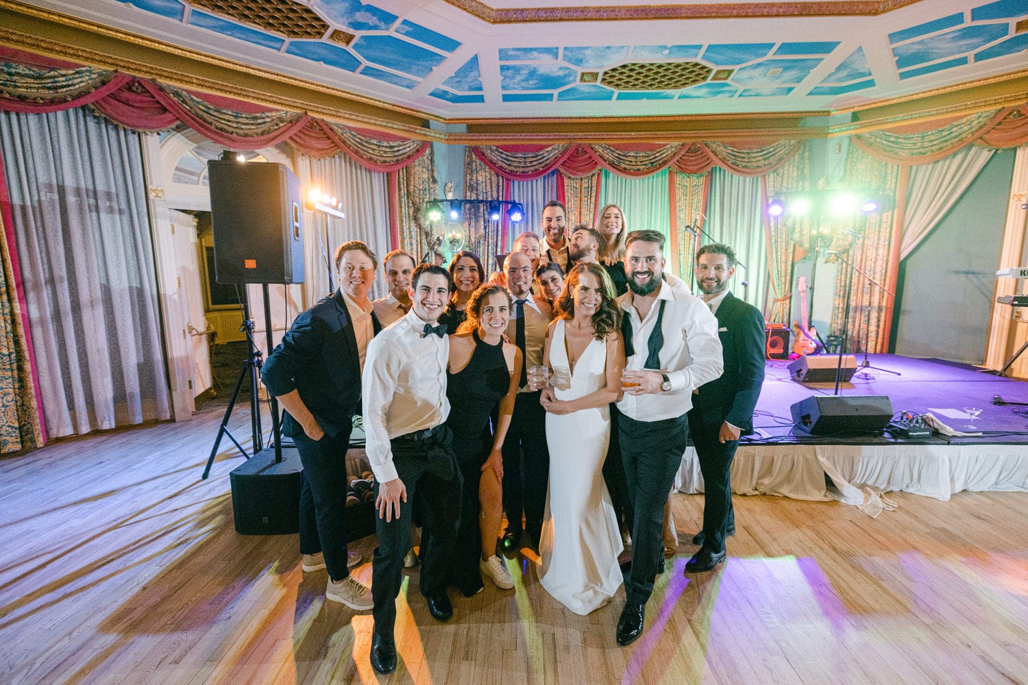 Group photo of joyful guests at a celebration in an elegant venue, with a bride in a white dress and friends in formal attire enjoying the moment.