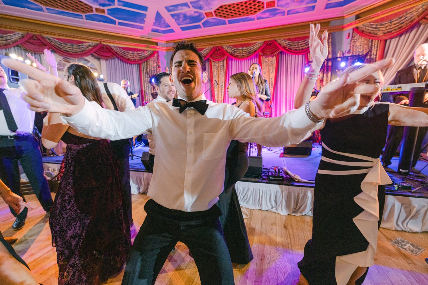 A lively man in a tuxedo enthusiastically dances among a crowd at a wedding reception, with a band performing in the background.