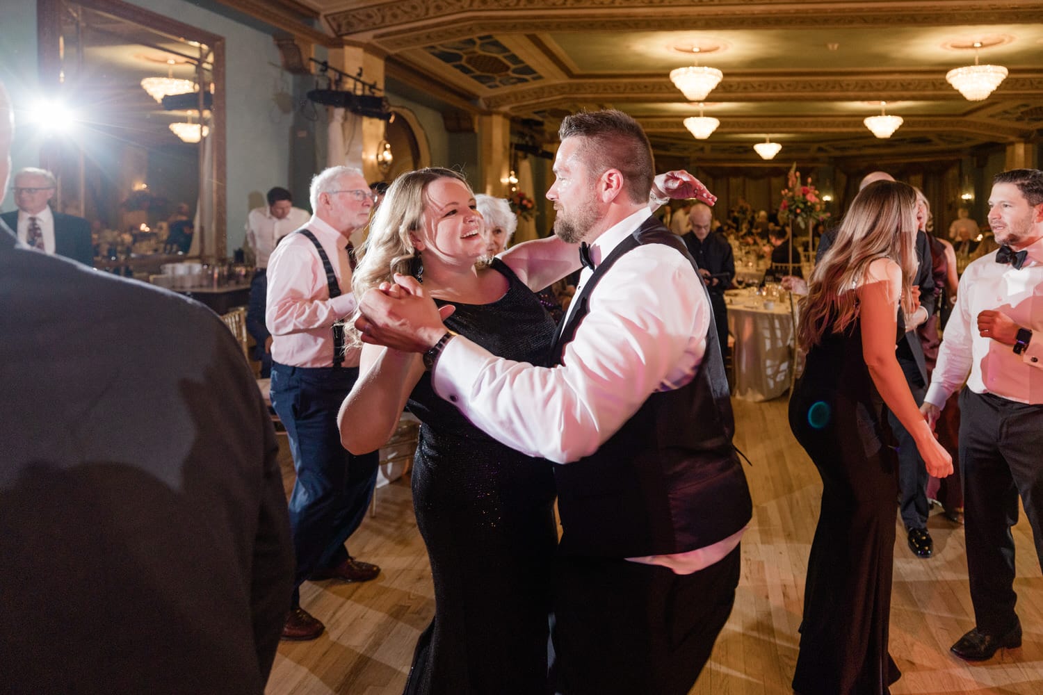 A couple joyfully dancing together at a lively celebration, surrounded by elegantly dressed guests in a beautifully decorated venue.