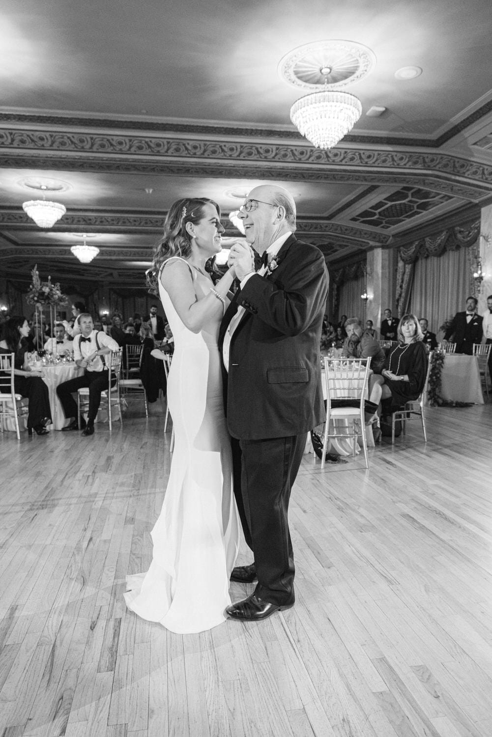 A joyful moment between a bride and her father during their dance at a wedding reception, captured in a beautifully lit venue with guests in the background.