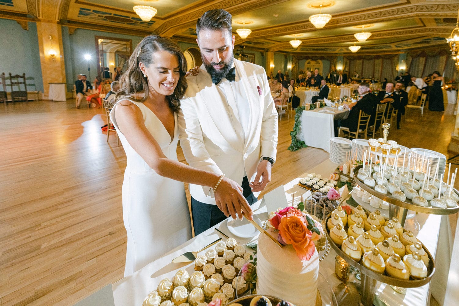 A happy couple in formal attire cutting a wedding cake adorned with flowers, surrounded by a variety of desserts in an elegant banquet hall.