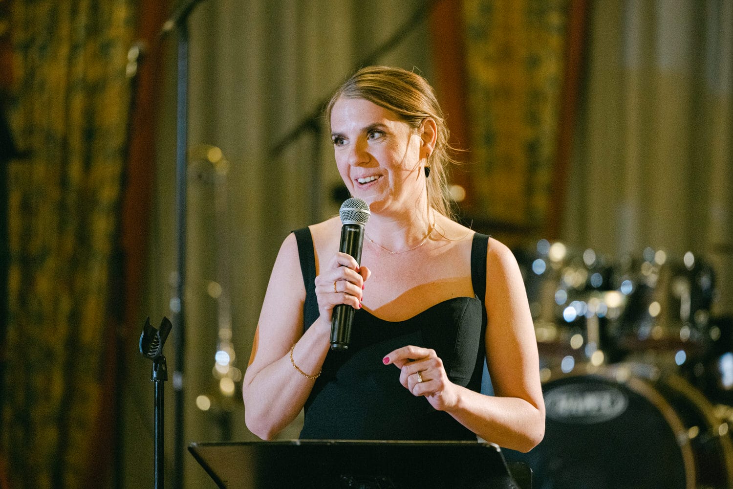 A woman in a black dress holding a microphone and smiling while speaking at a microphone stand, with a band setup in the background.