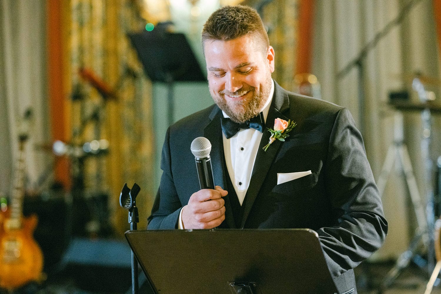 A smiling man in a tuxedo holds a microphone while giving a toast at a wedding, with a music setup in the background.