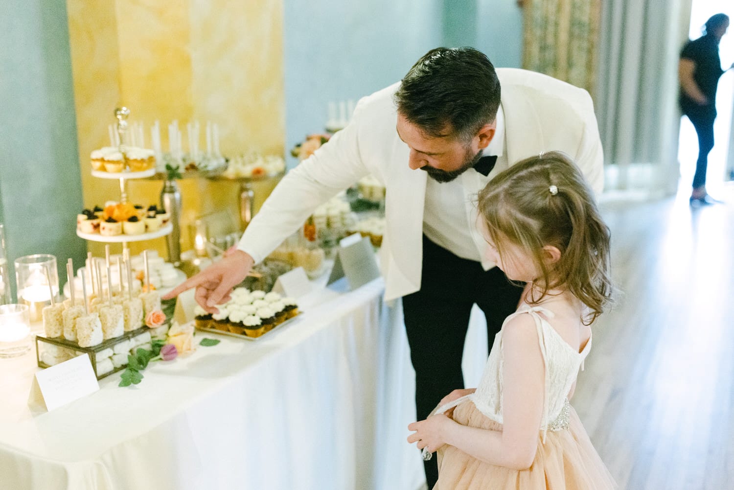 A man in a white suit interacts with a young girl in a dress as they explore a dessert table filled with various treats and desserts.