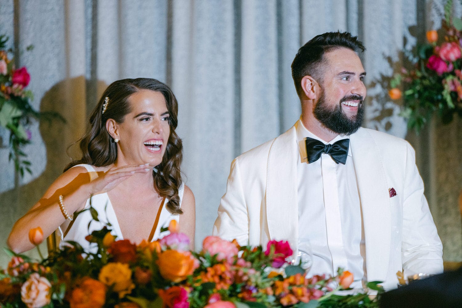 A happy couple during their wedding reception, laughing and smiling at each other with a vibrant floral arrangement in the foreground.